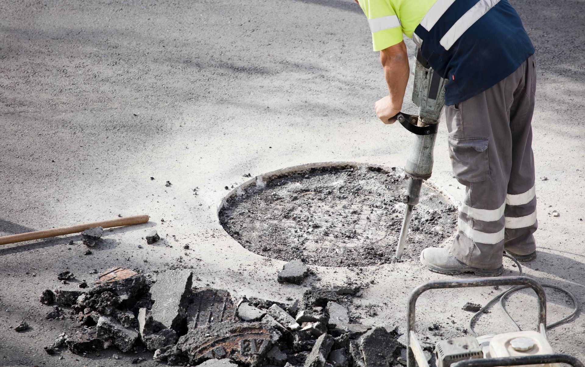 Person in safety vest and reflective pants using a jackhammer to break up asphalt around a manhole.