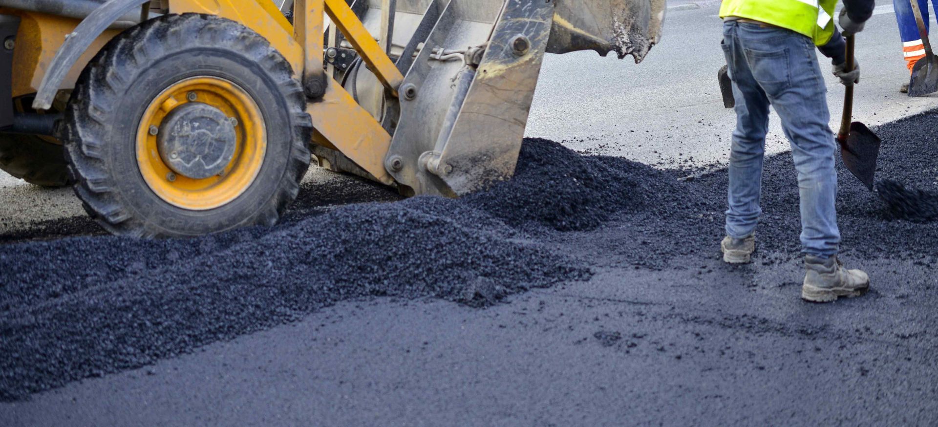 A yellow construction vehicle and a worker with a shovel laying asphalt on a road.