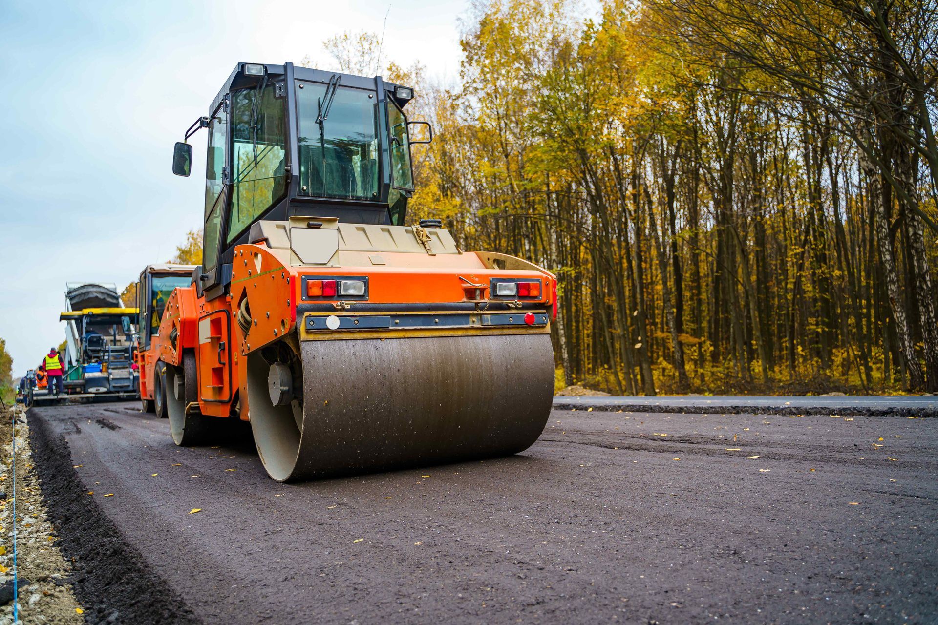 Road roller compacting fresh asphalt on a road, with trees in the background.