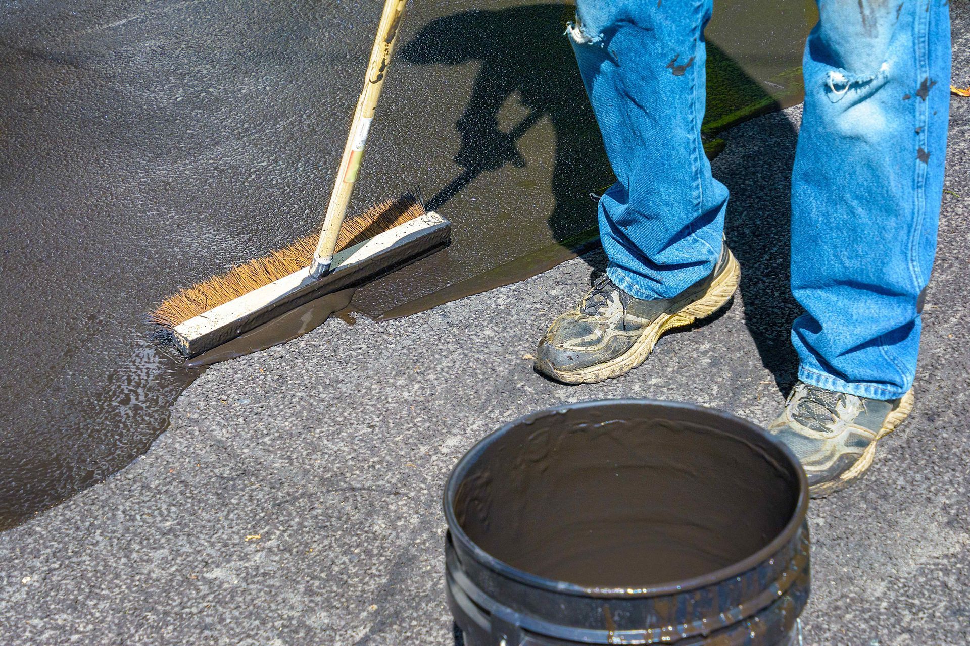 Person applying black sealant to asphalt with a squeegee; bucket of sealant in foreground.
