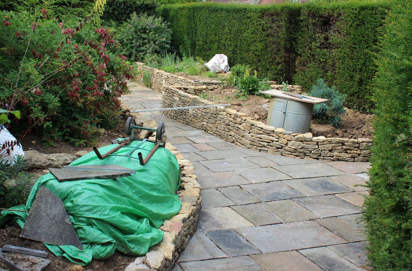 Stone path winds through garden with retaining walls, hedges, and construction materials.