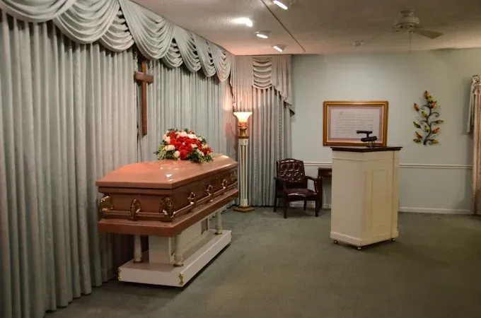 A casket with floral arrangement in a funeral home chapel; curtains, crucifix, lectern, and chair.