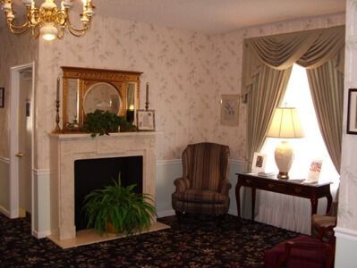 Living room with fireplace, armchair, and window with drapes. Includes lamp, table, and ornate wallpaper.