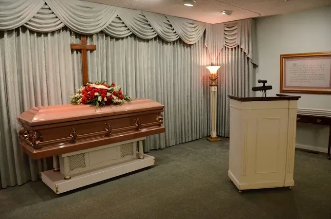 Funeral home chapel. Casket with floral arrangement, wooden cross, podium, light-colored drapes.