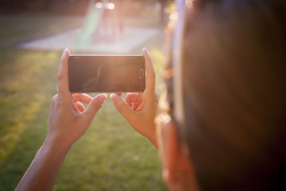 Person holding a smartphone, taking a photo of a playground in a sunny outdoor setting.