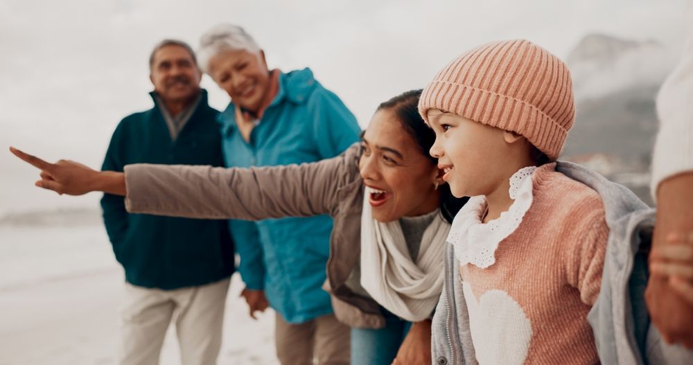 Family on beach, woman points, child smiles, grandparents watch, overcast sky.