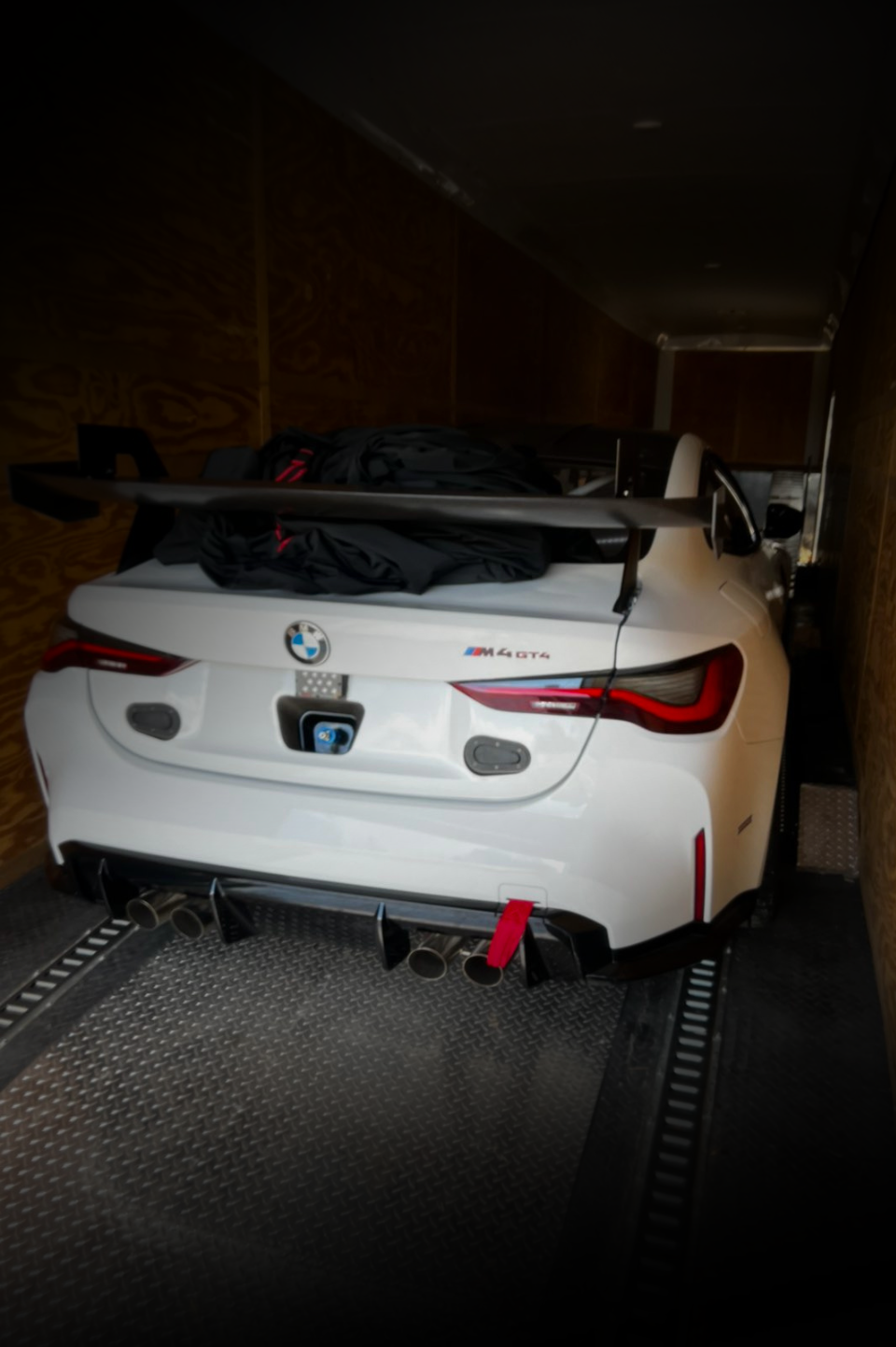 A white BMW M4 sports car with a large rear wing and a red tow strap, parked inside an enclosed cargo trailer.