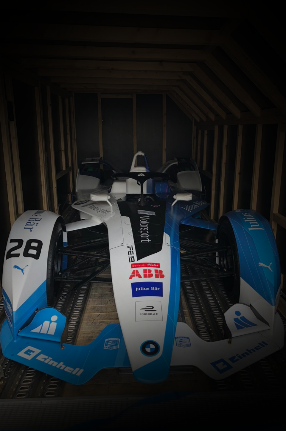 A BMW Formula E race car, numbered 28 with blue and white branding, is parked inside a wooden-framed trailer.