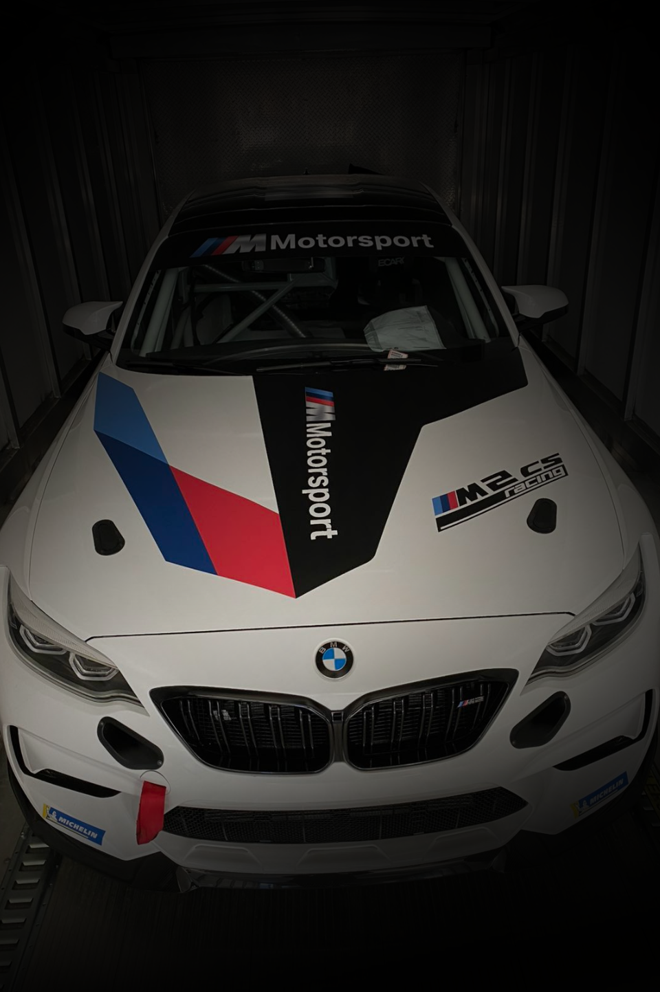 A white BMW M2 CS Racing car with motorsport livery, parked inside a trailer.