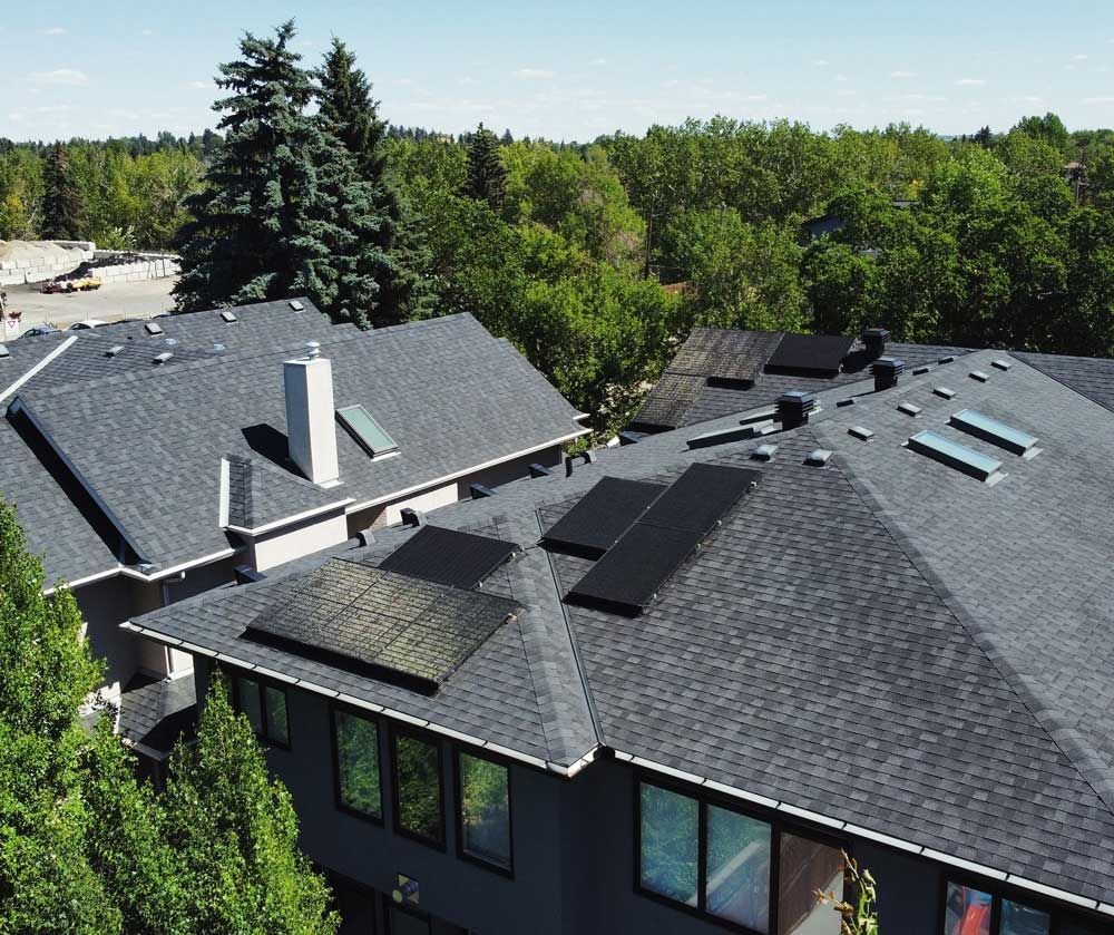 Black solar panels on a gray roof, surrounded by trees.