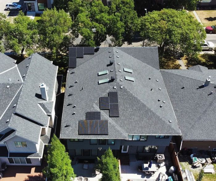 Aerial view of a dark gray shingled roof with solar panels installed, surrounded by trees and neighboring houses.