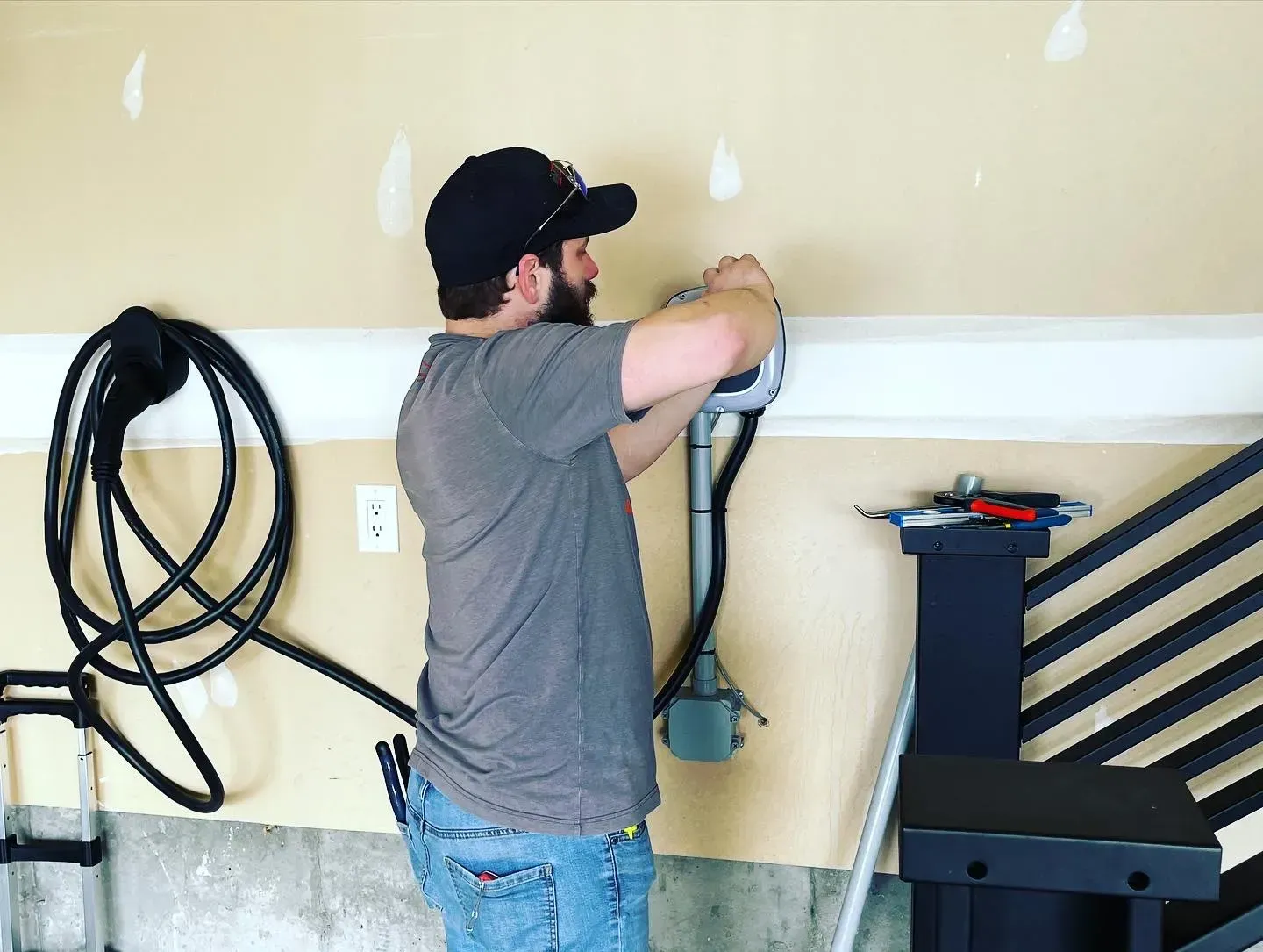 Man in gray shirt installing an electric car charger on a beige garage wall.