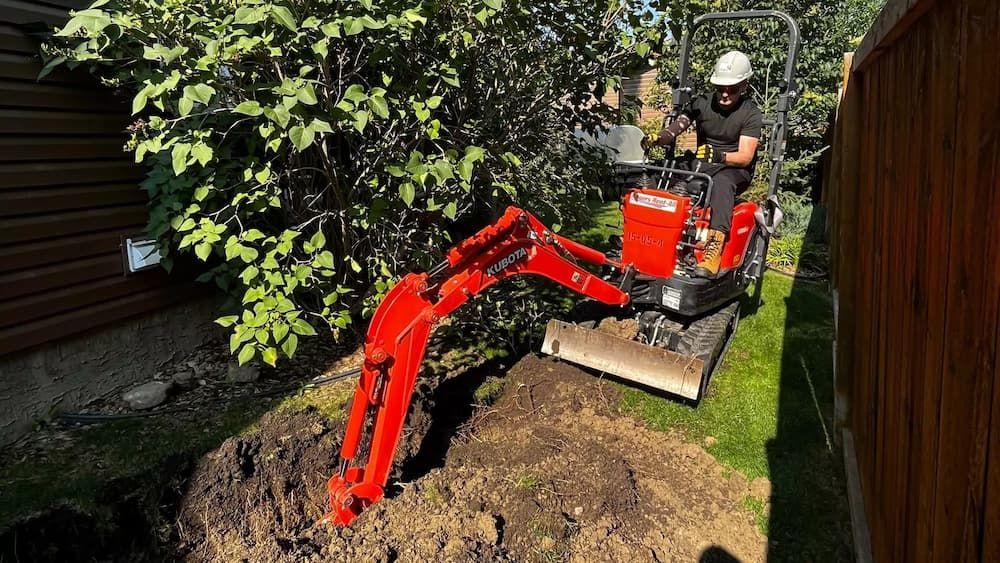 Person operating a red mini-excavator, digging a trench in a grassy backyard.