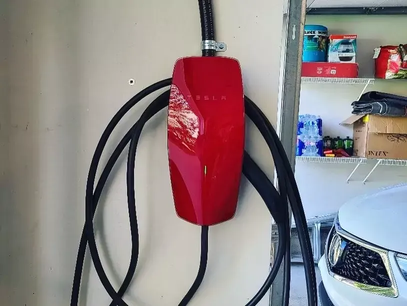 Aerial view of a house with solar panels on a dark roof, surrounded by trees and green grass. A vehicle is on the driveway.