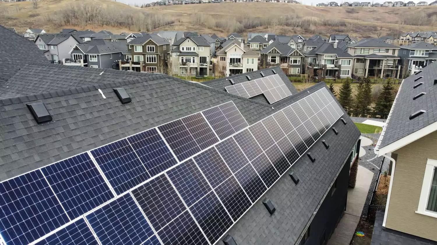Solar panels on a dark roof in a residential neighborhood with gray rooftops and trees.