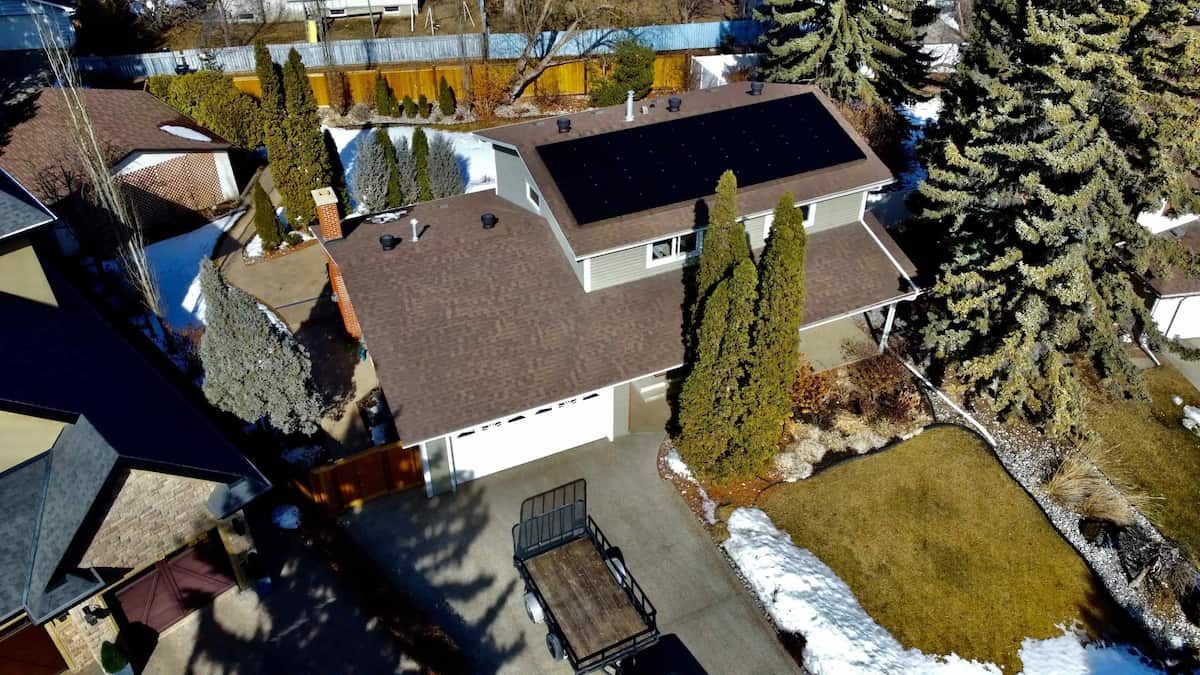 Aerial view of a house with solar panels on the roof. Brown roof, white garage, snowy yard, and green trees.