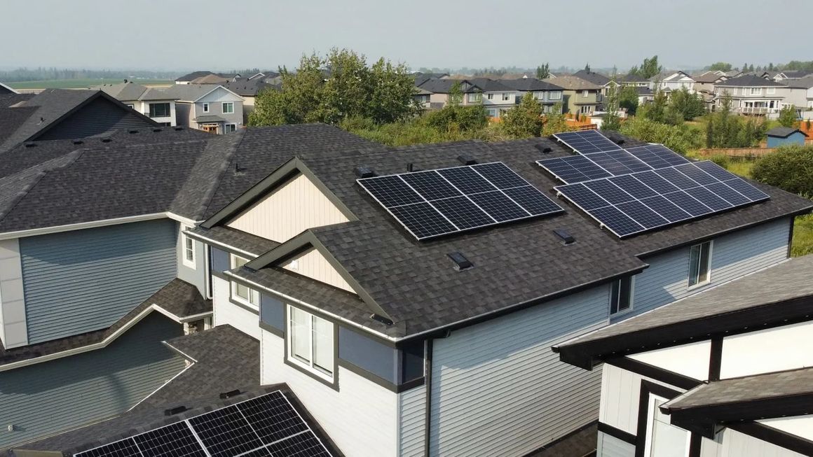 Solar panels on a residential rooftop in a suburban neighborhood. Blue panels on dark shingle roof.