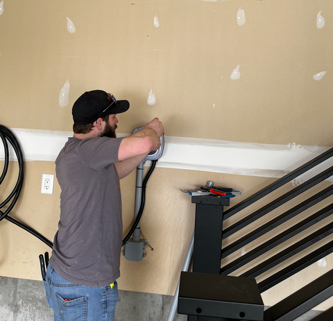Man in cap works on electrical wiring on a beige wall in a garage.