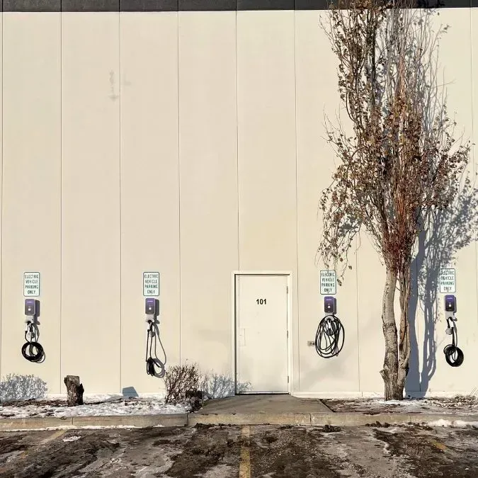 Exterior wall with three EV chargers, a door, and a small tree, snowy parking lot.