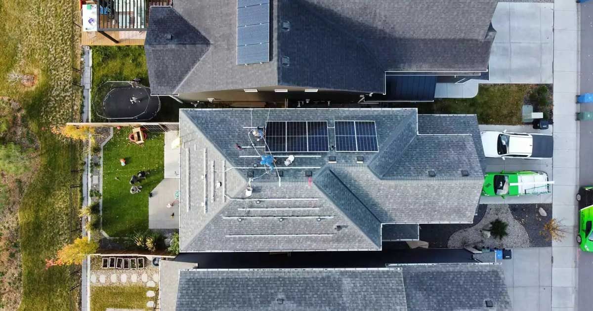 Overhead view of rooftops with solar panels; house with green lawn, driveway, and parked truck.