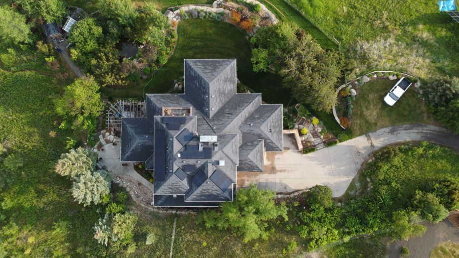 Aerial view of a large house with a complex, dark gray roof, surrounded by green trees and grass.