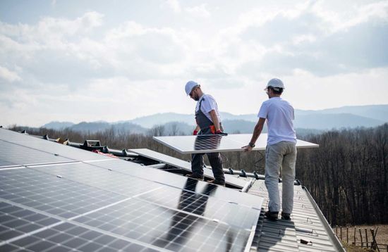 Two workers installing a solar panel on a rooftop with mountains in the background.
