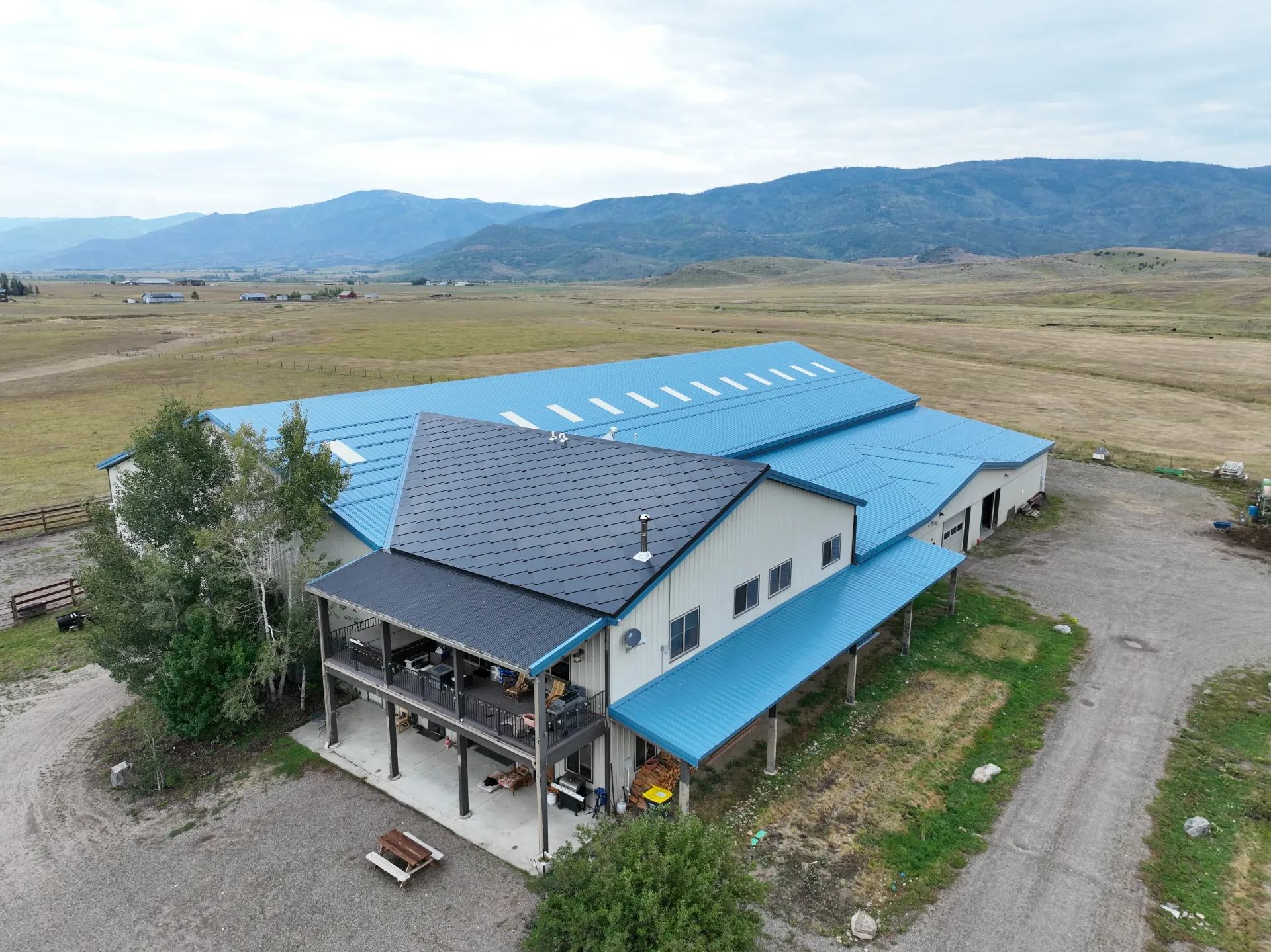 Aerial view of a dark gray shingled roof with solar panels installed, surrounded by trees and neighboring houses.