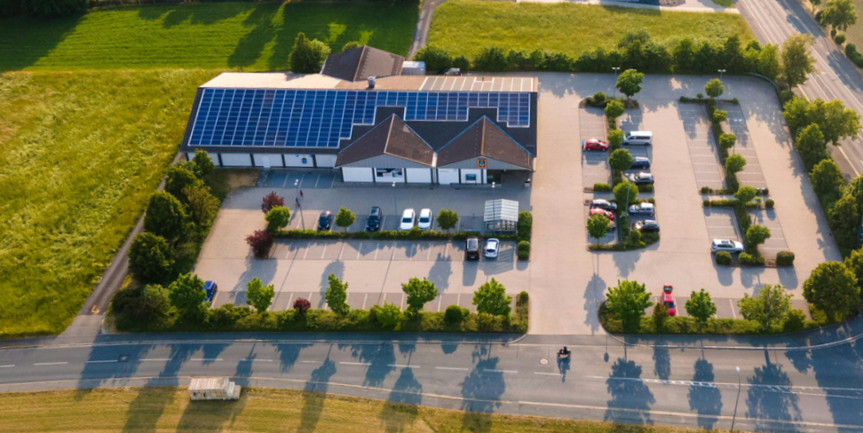 Aerial view of a building with solar panels, a parking lot, and surrounding trees and fields.