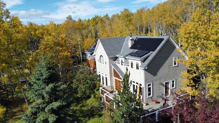 Two-story house with gray roof, solar panels, surrounded by autumn trees with yellow and green foliage.