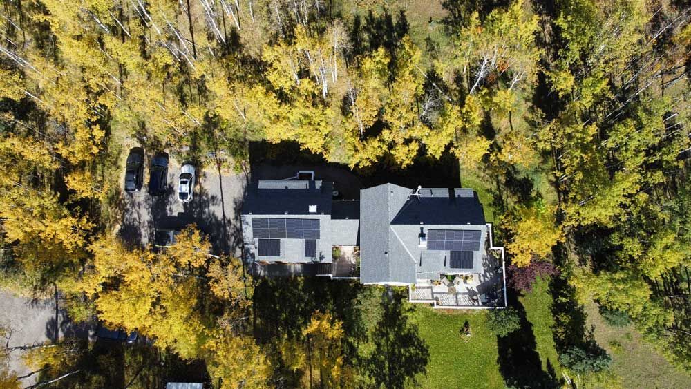 Aerial view of a house with solar panels, surrounded by trees with yellow autumn foliage and a parked car.