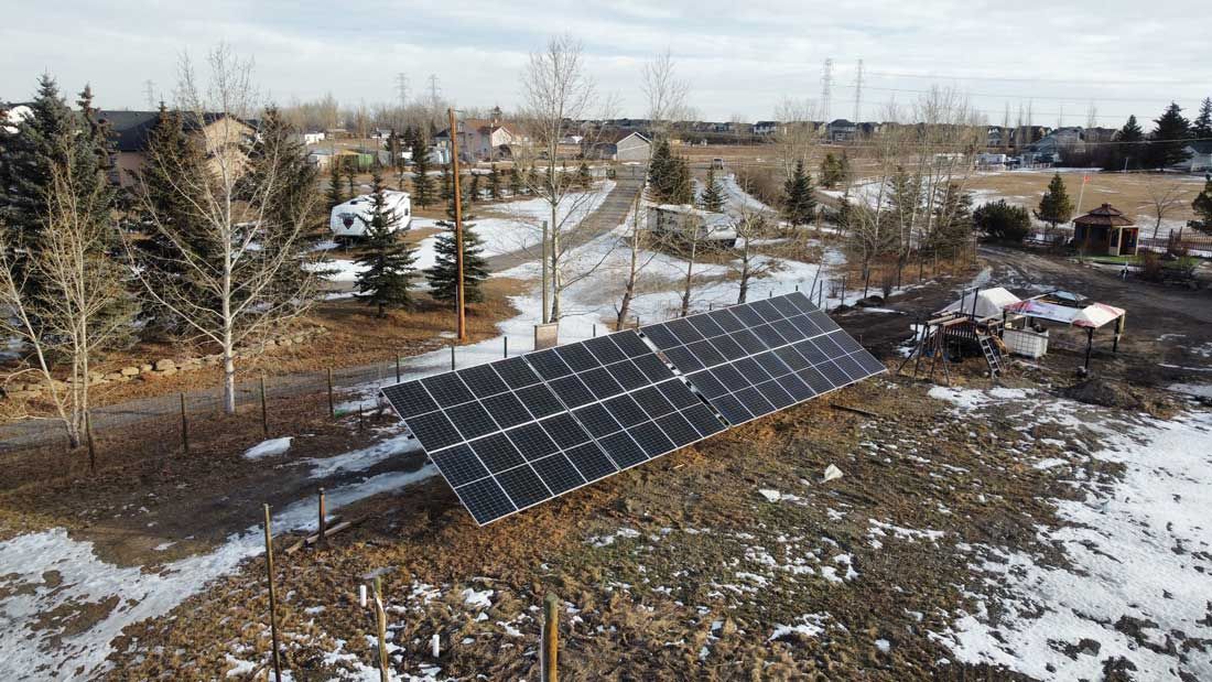 Solar panels on a slight hill, surrounded by snow and trees in a rural setting.