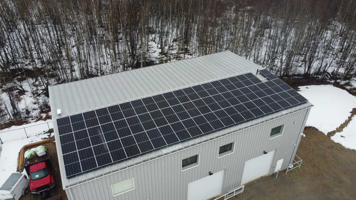 Solar panels on a metal roof in winter; building with a snow-covered surrounding and trees.