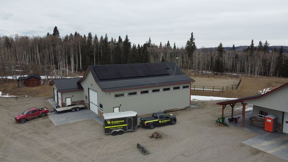 Gray building with solar panels, vehicles, and a trailer on a gravel lot, with a forest backdrop.