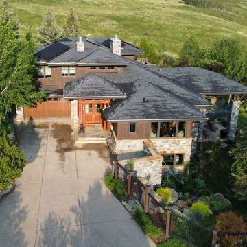 Brown house with a dark gray roof, set against a green hillside. Concrete driveway, stone and wood accents.
