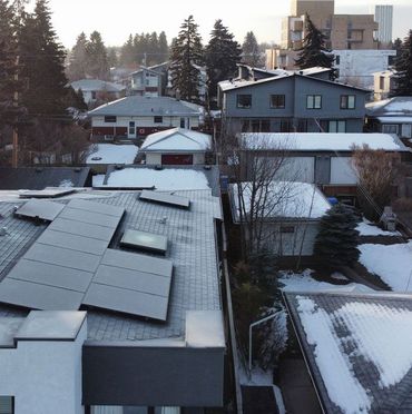 Snowy rooftops of houses with solar panels, trees, and buildings in the background.