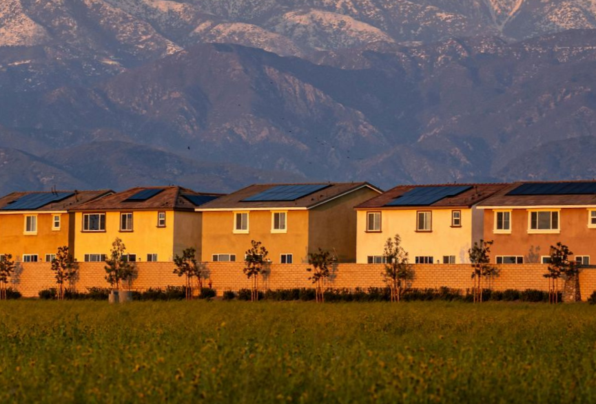 Row of houses with solar panels, green field, and mountains in the background.