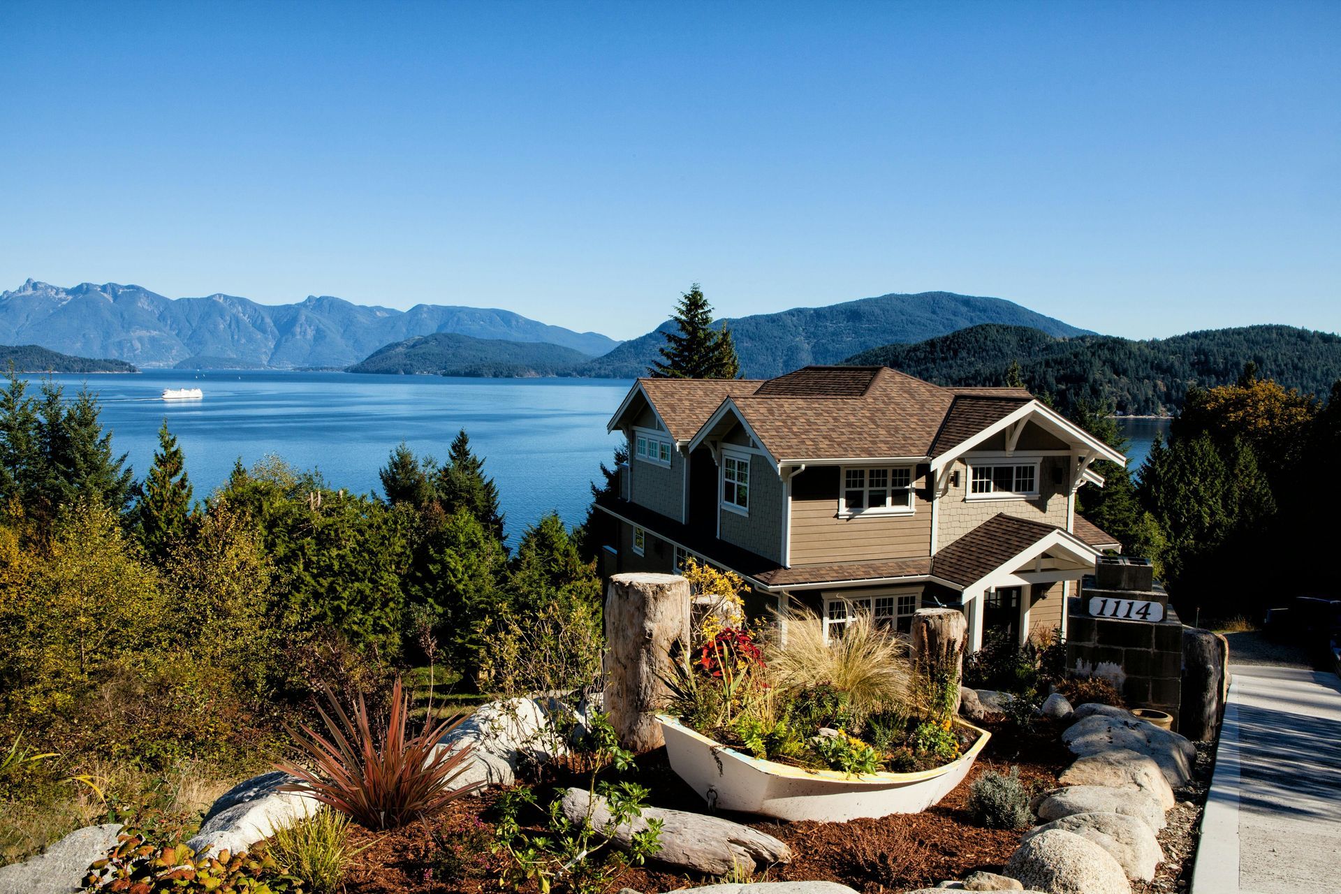 House overlooking a lake with mountains under a blue sky, surrounded by trees and a garden.