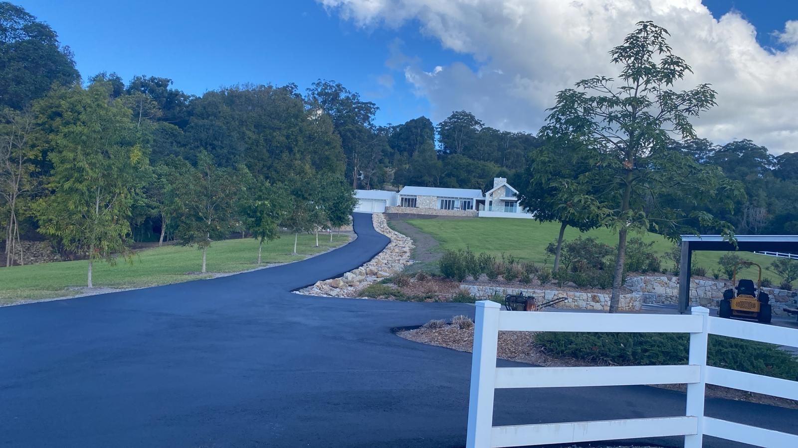 A blue asphalt roller compacting freshly laid asphalt driveway in front of a house and shed.