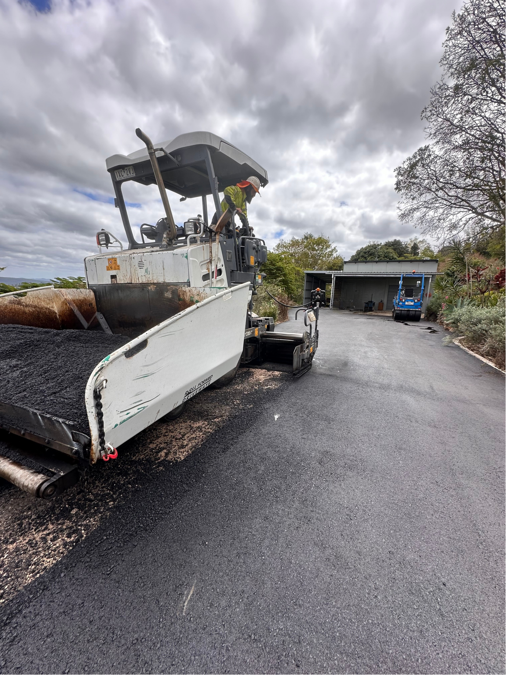A Man Is Standing Next To A Machine That Is Laying Asphalt On A Road — Roll Formed Driveways in Gympie, QLD