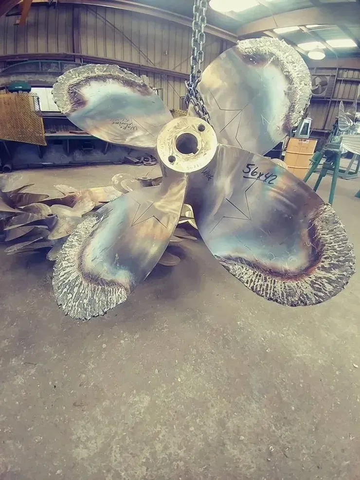 A large metal propeller is hanging from chains in a warehouse.