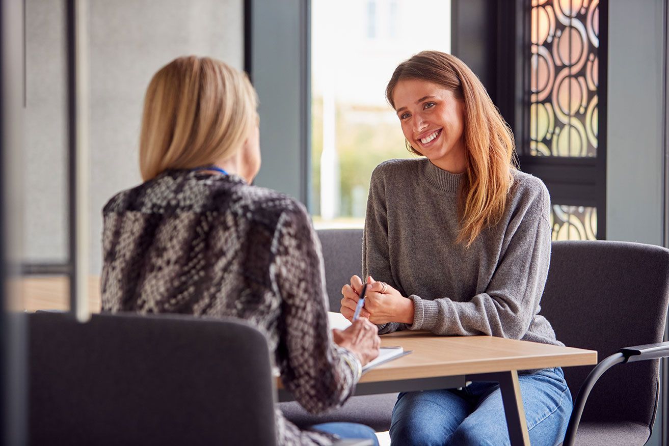 Two people talking at a table in a bright office, one smiling and holding a pen