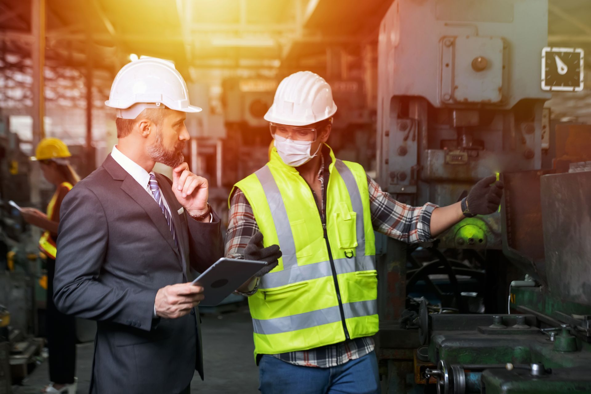 Two men in hard hats and safety vests are standing in a factory looking at a tablet.