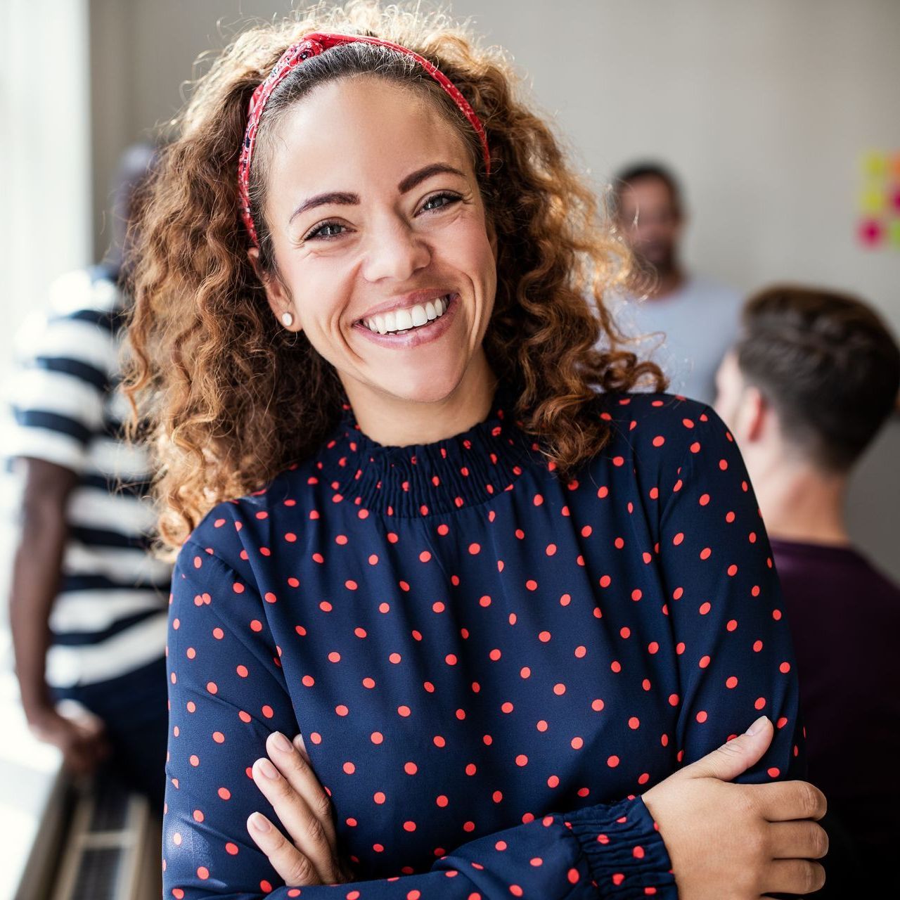 Smiling woman with curly hair, red headband, and blue polka-dot shirt, arms crossed.