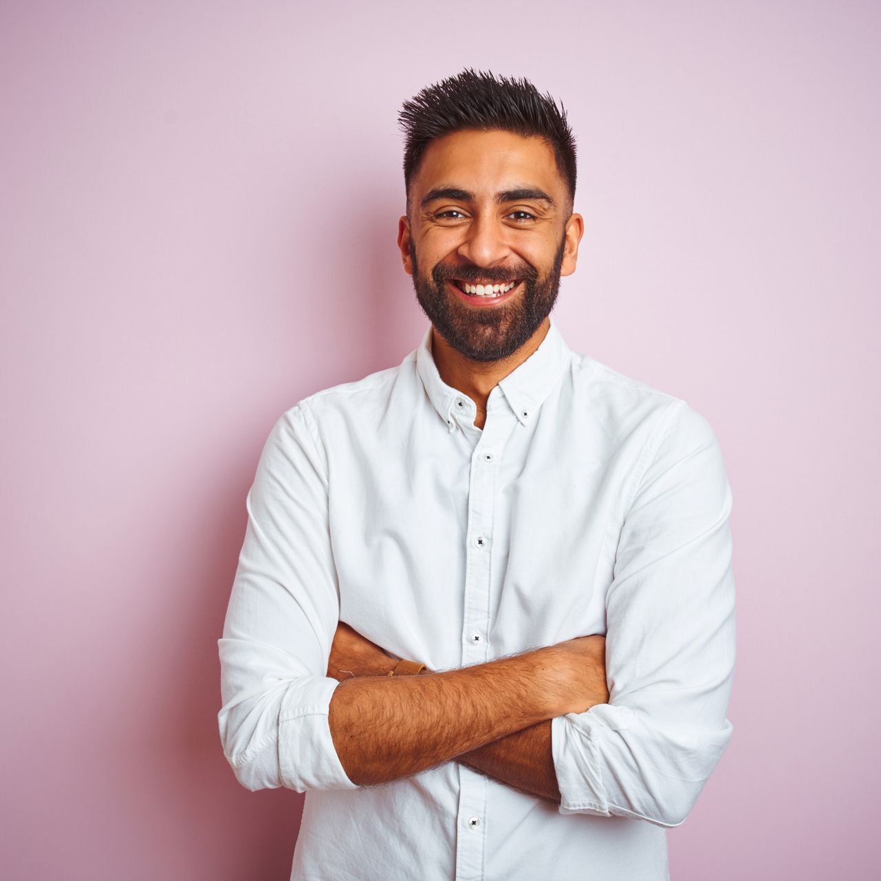 Man with a dark beard and a white button-up shirt smiles with arms crossed, standing in front of a pink background.
