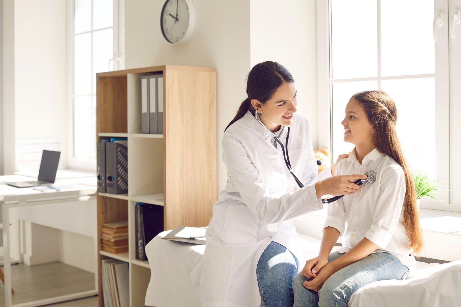 A female doctor examines a smiling young girl with a stethoscope in a sunlit medical office.