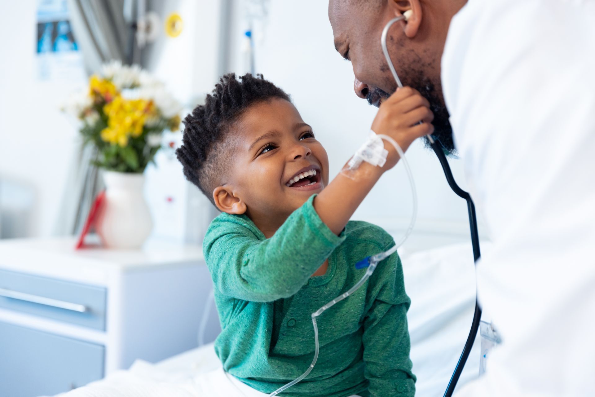 Smiling young boy in a hospital bed playfully interacts with a doctor wearing a stethoscope. They are both Black.