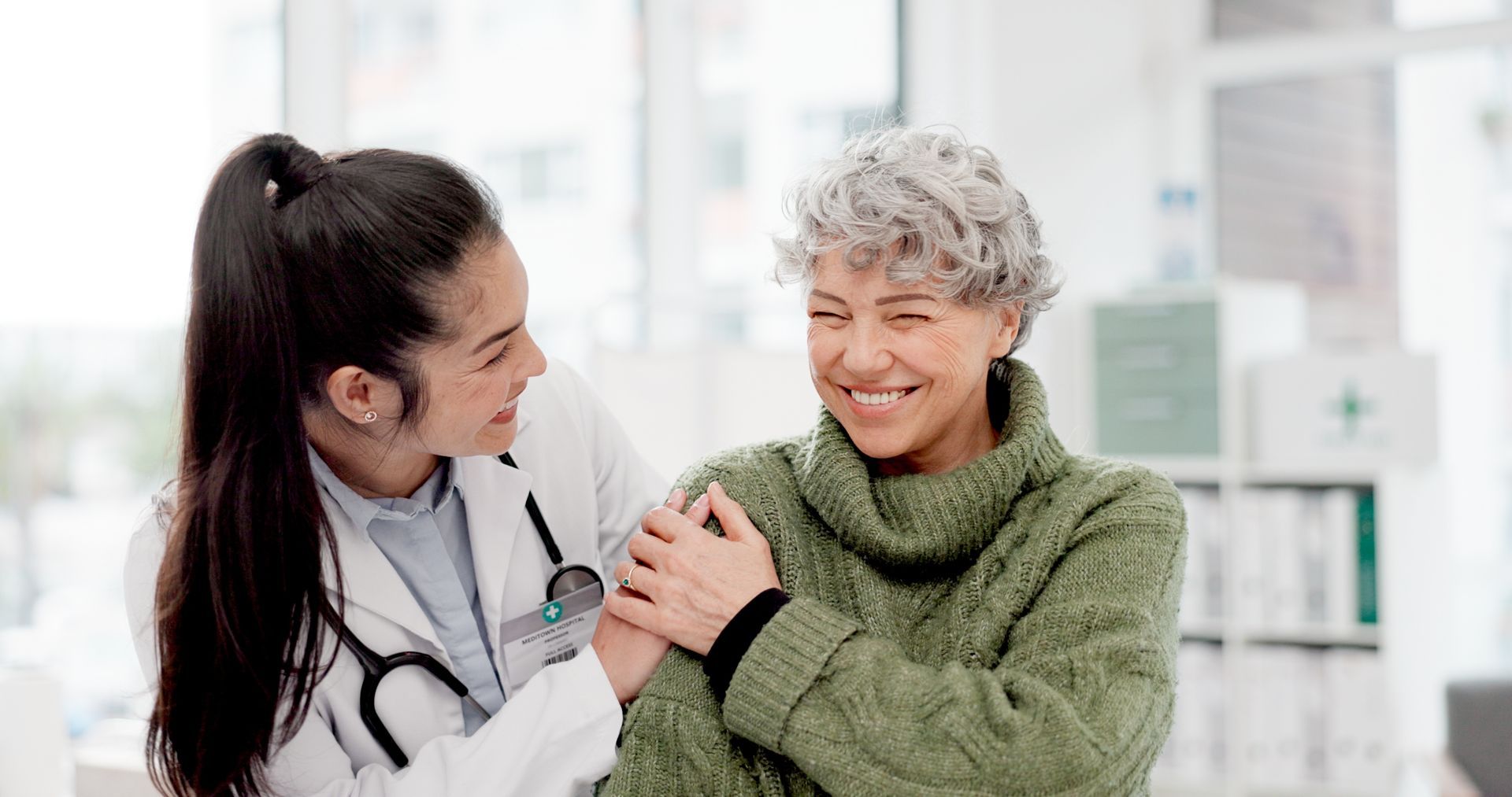 A young doctor with a stethoscope puts her arm around an older woman in a green sweater, both smiling in a medical office.