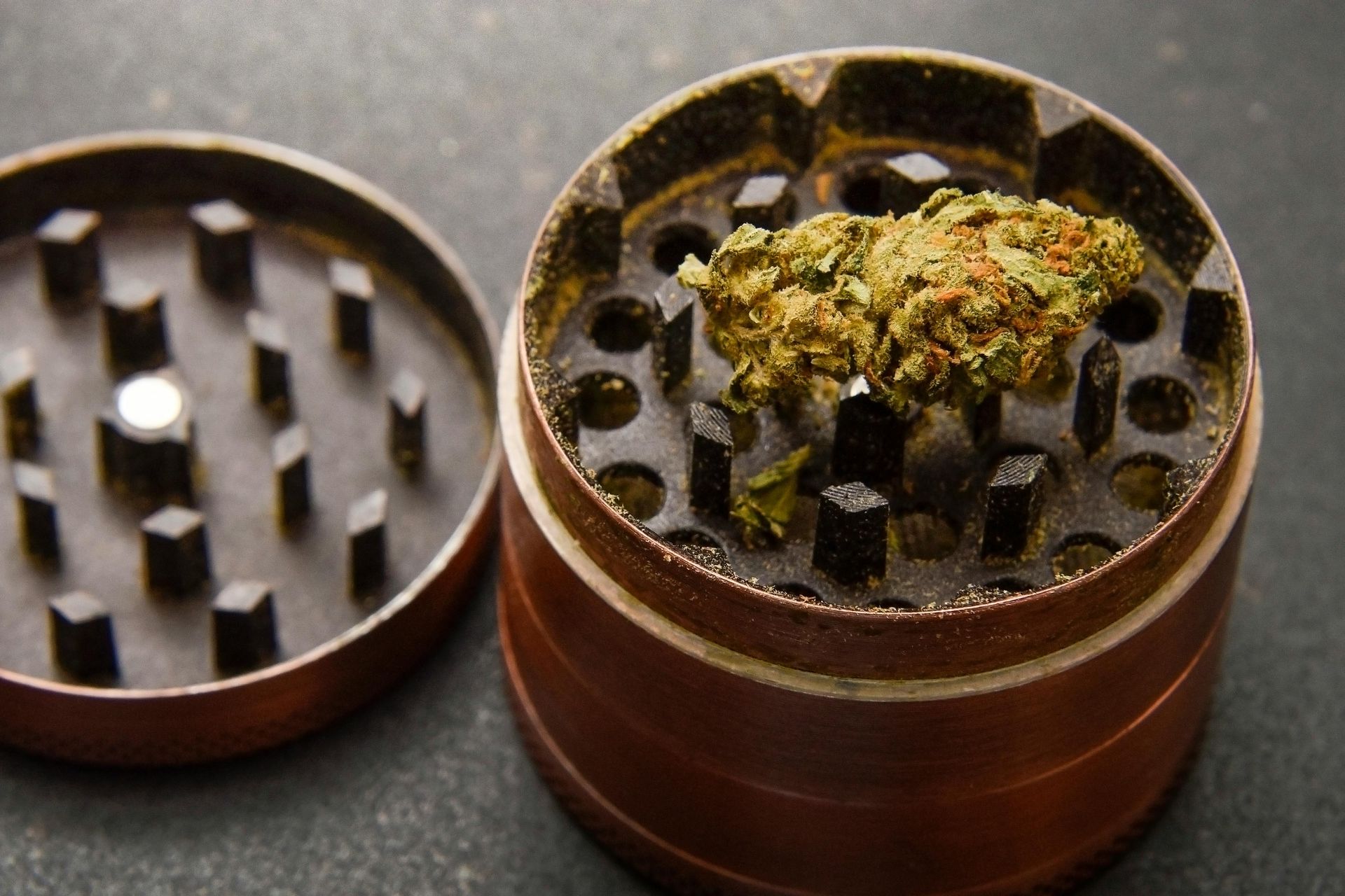 A copper-colored metal herb grinder open with a small cannabis bud resting on its teeth, set against a dark background.