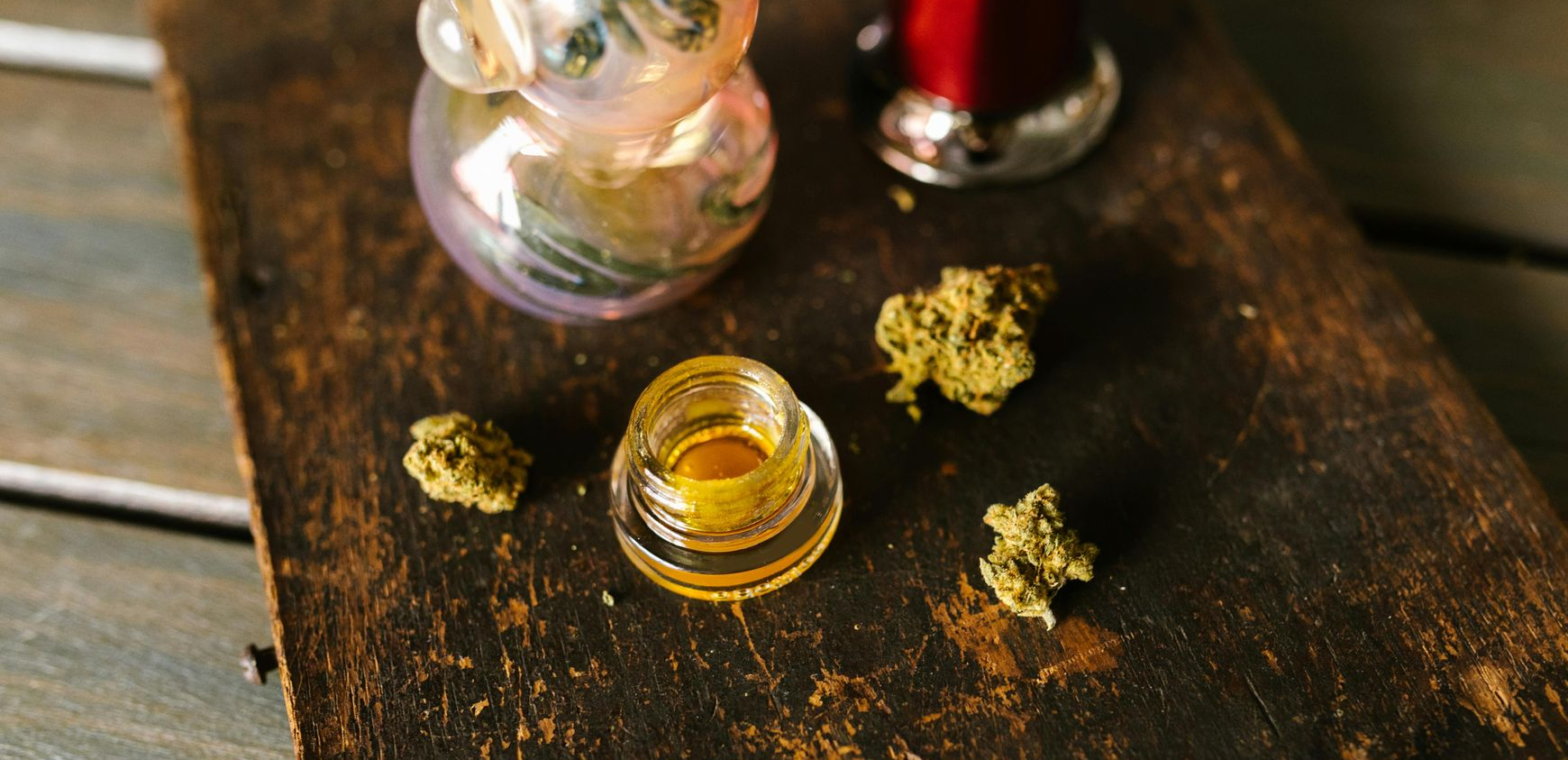 A glass pipe, a jar of cannabis concentrate, and small cannabis flower buds arranged on a dark, rustic wooden surface.