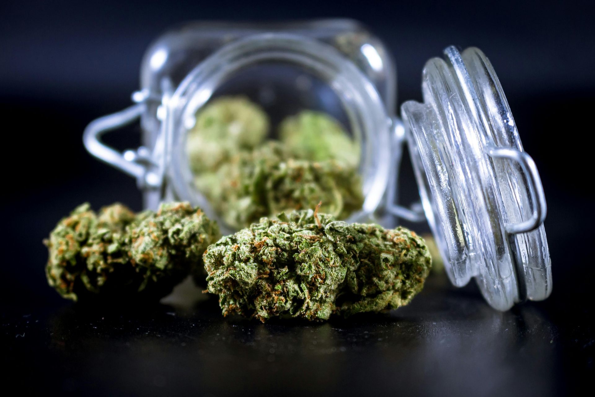 Green cannabis buds sit on a black surface next to a glass jar with an open lid.